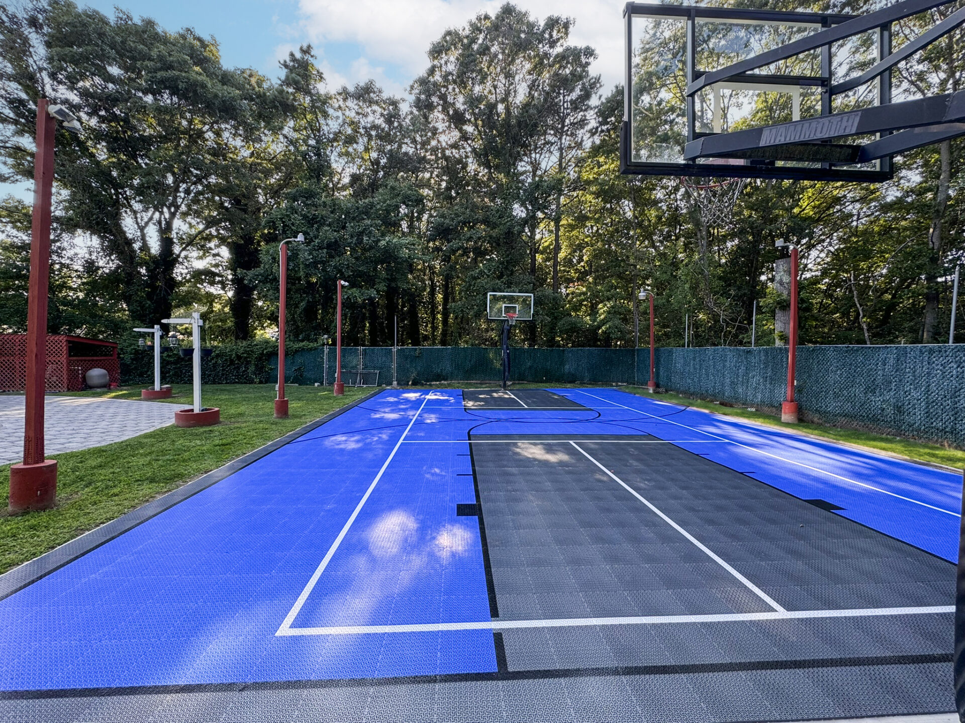 Outdoor basketball court with blue and black surfaces, surrounded by a fence and trees, under a partly cloudy sky.