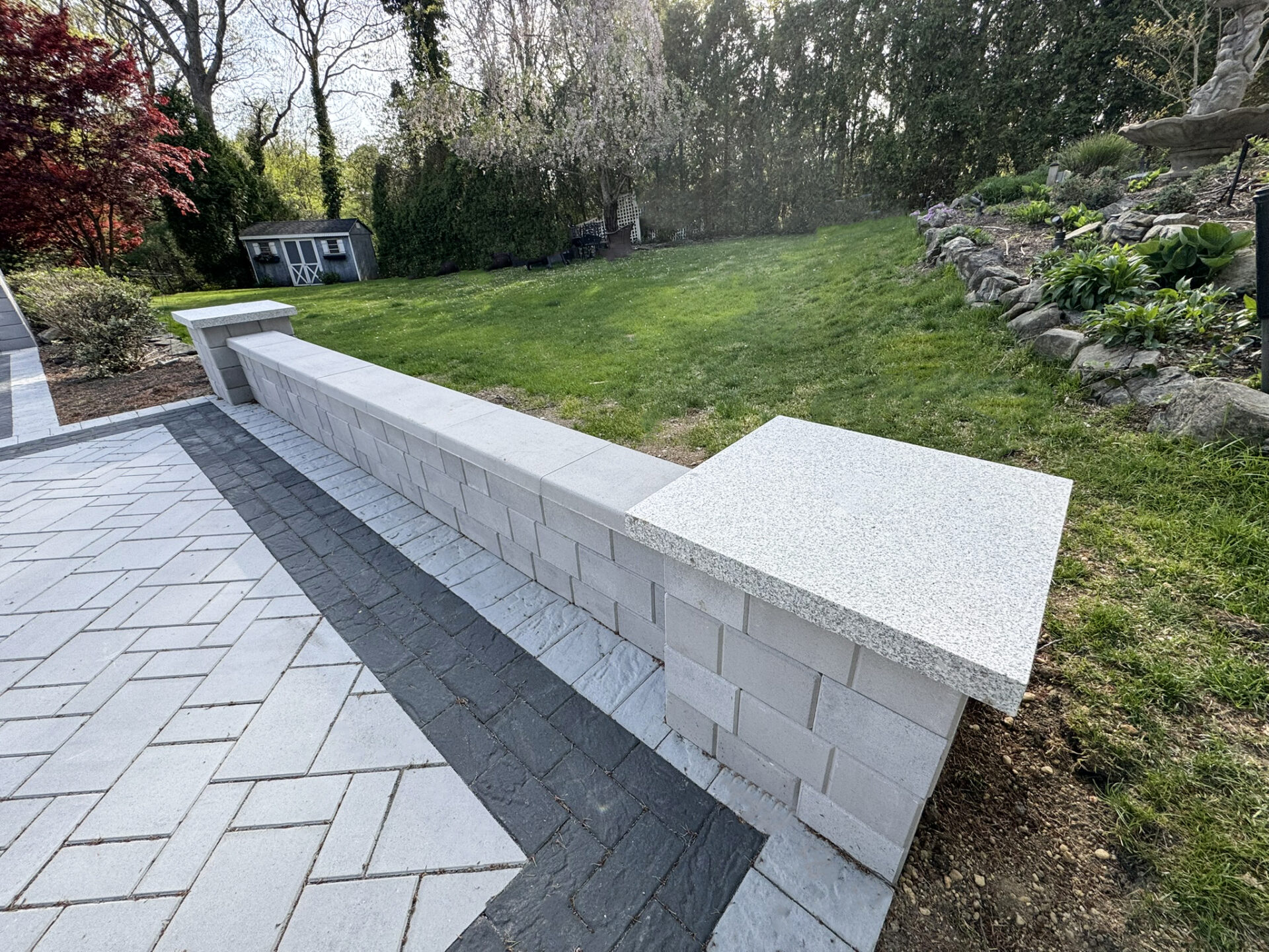 A low concrete block wall with stone caps separates a paved patio area from a grassy backyard with landscaping and a shed in the background.