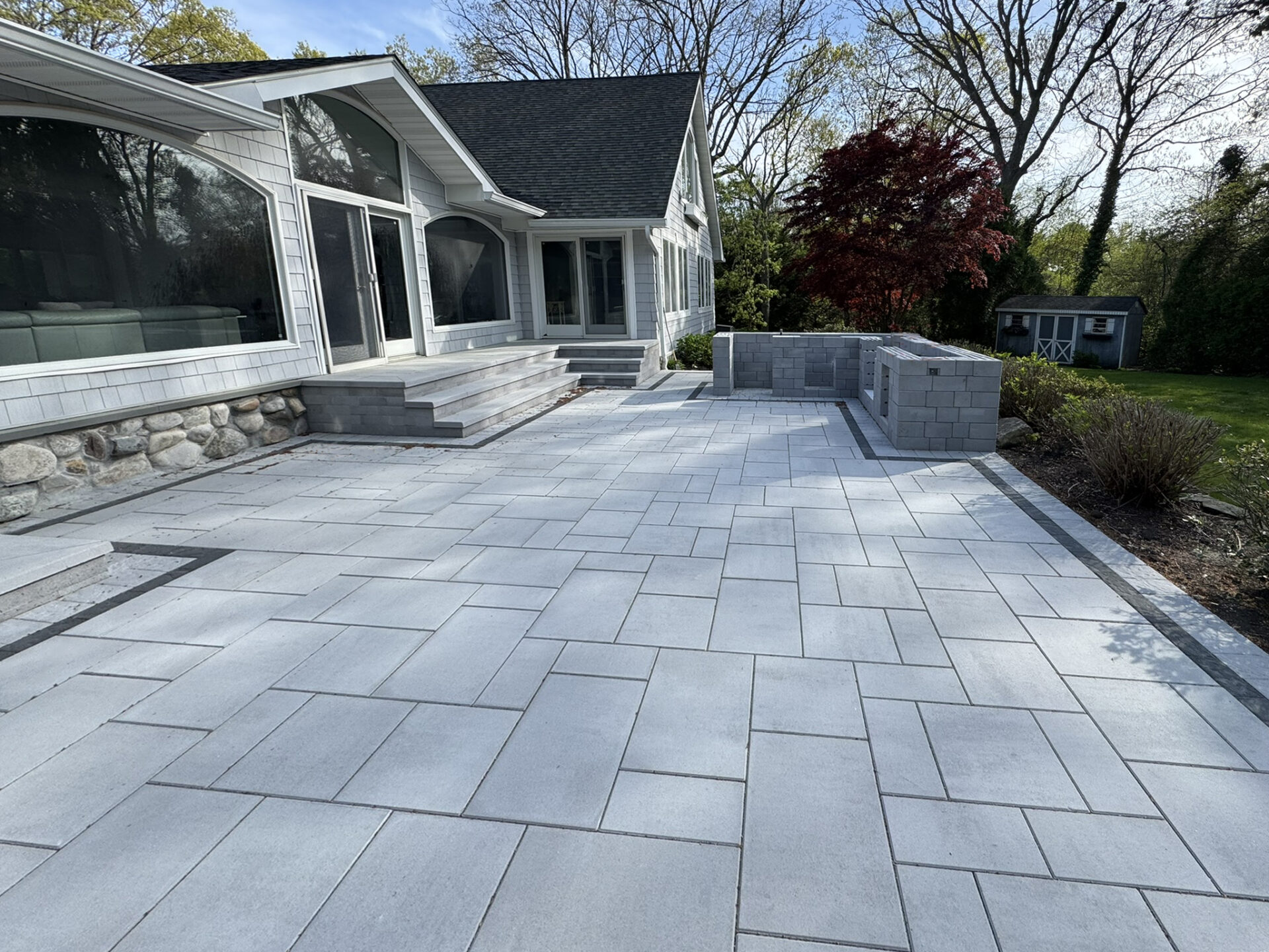 A modern house with large windows and stone accents features a spacious outdoor patio with light gray pavers and a low stone wall, surrounded by trees and a small lawn.