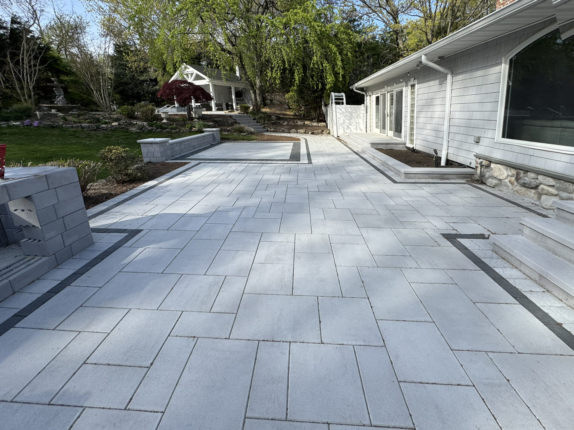 A large stone patio with light gray pavers and dark border accents, adjacent to a house with white siding and surrounded by landscaped trees and shrubs.