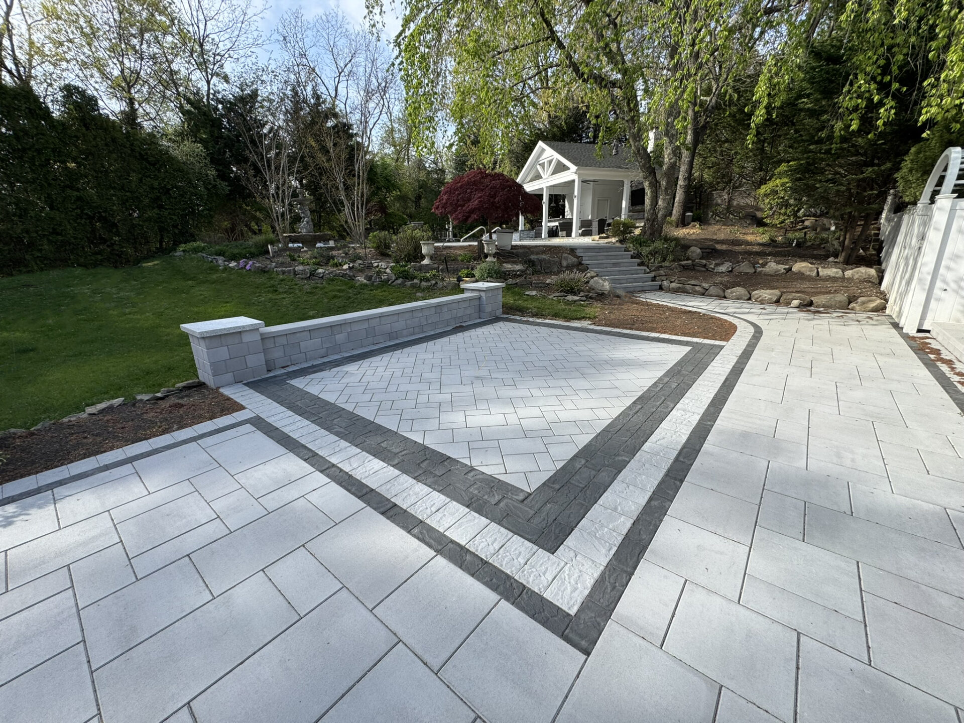 A backyard patio with light gray pavers, bordered by dark gray pavers, featuring a small retaining wall, garden, and a white gazebo in the background.