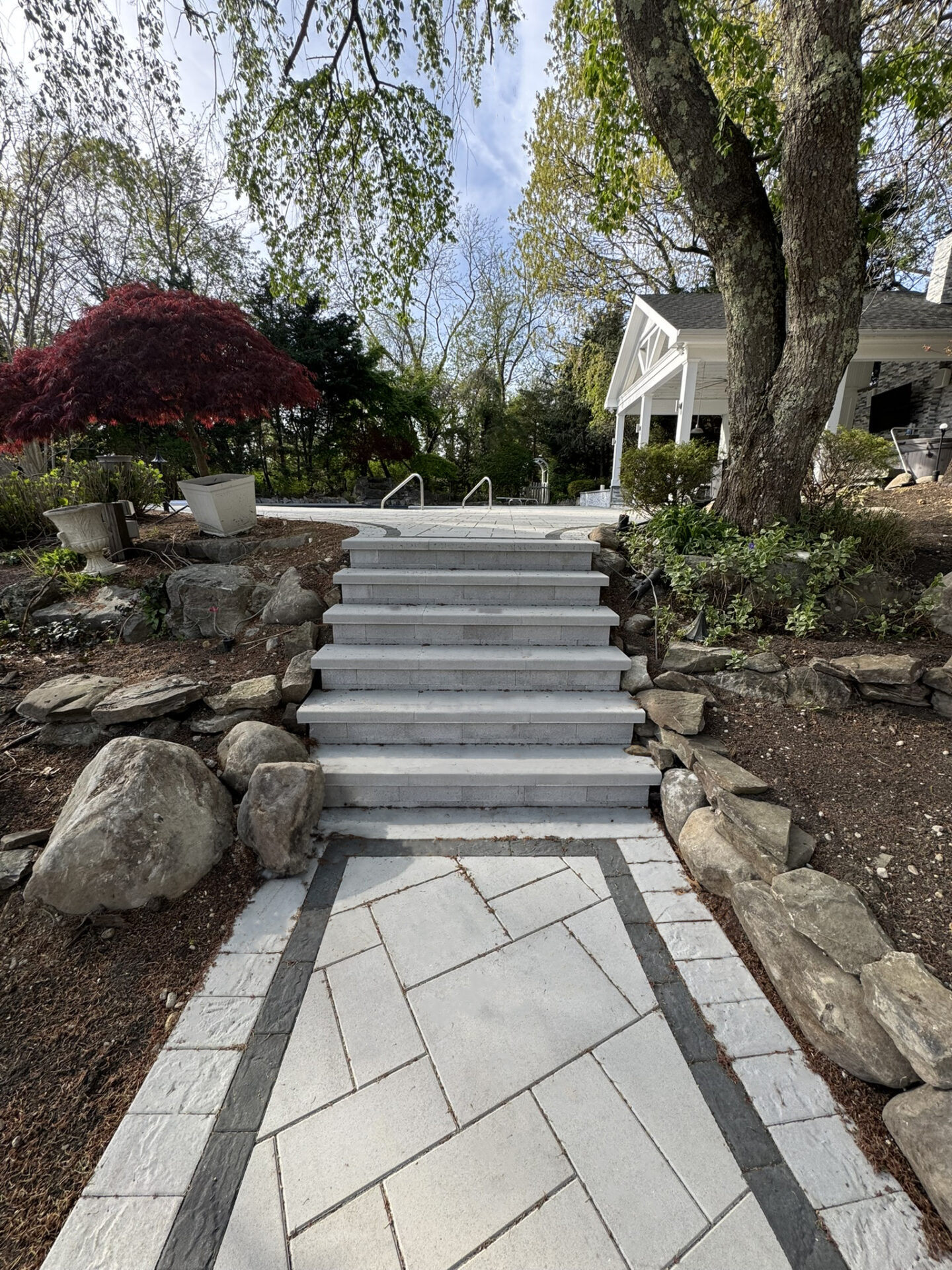 Stone steps lead up from a paved walkway between rocks and trees toward a white house with a porch in a landscaped garden.