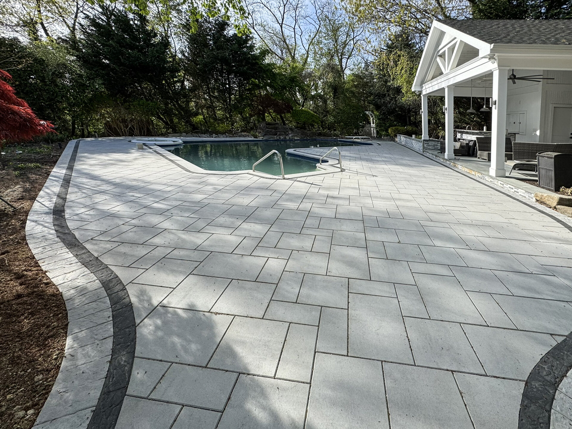 Rectangular pool with metal handrails, surrounded by light gray paver stones and a curved dark border, adjacent to a covered outdoor seating area and trees in the background.