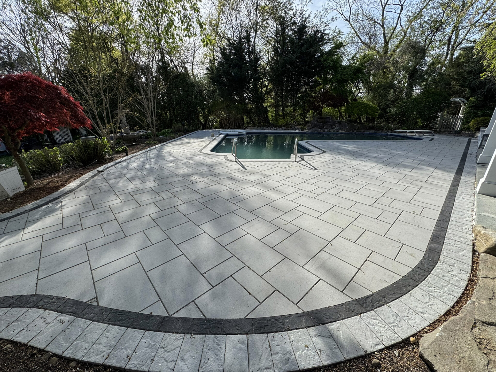 A small backyard swimming pool is surrounded by new light gray stone pavers with a darker border, set in a landscaped area with trees and shrubs.