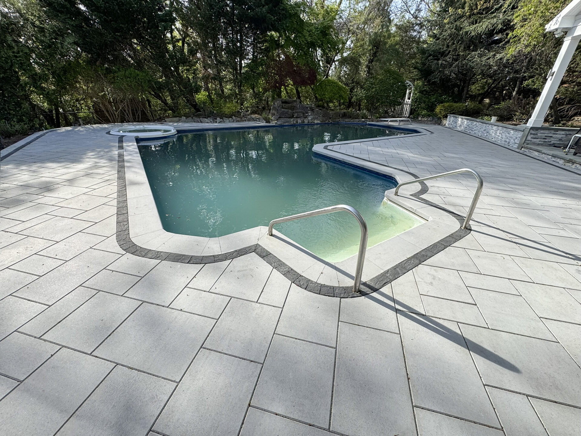 An in-ground swimming pool with light-colored tile decking, metal handrails, and a nearby hot tub, surrounded by trees and greenery.