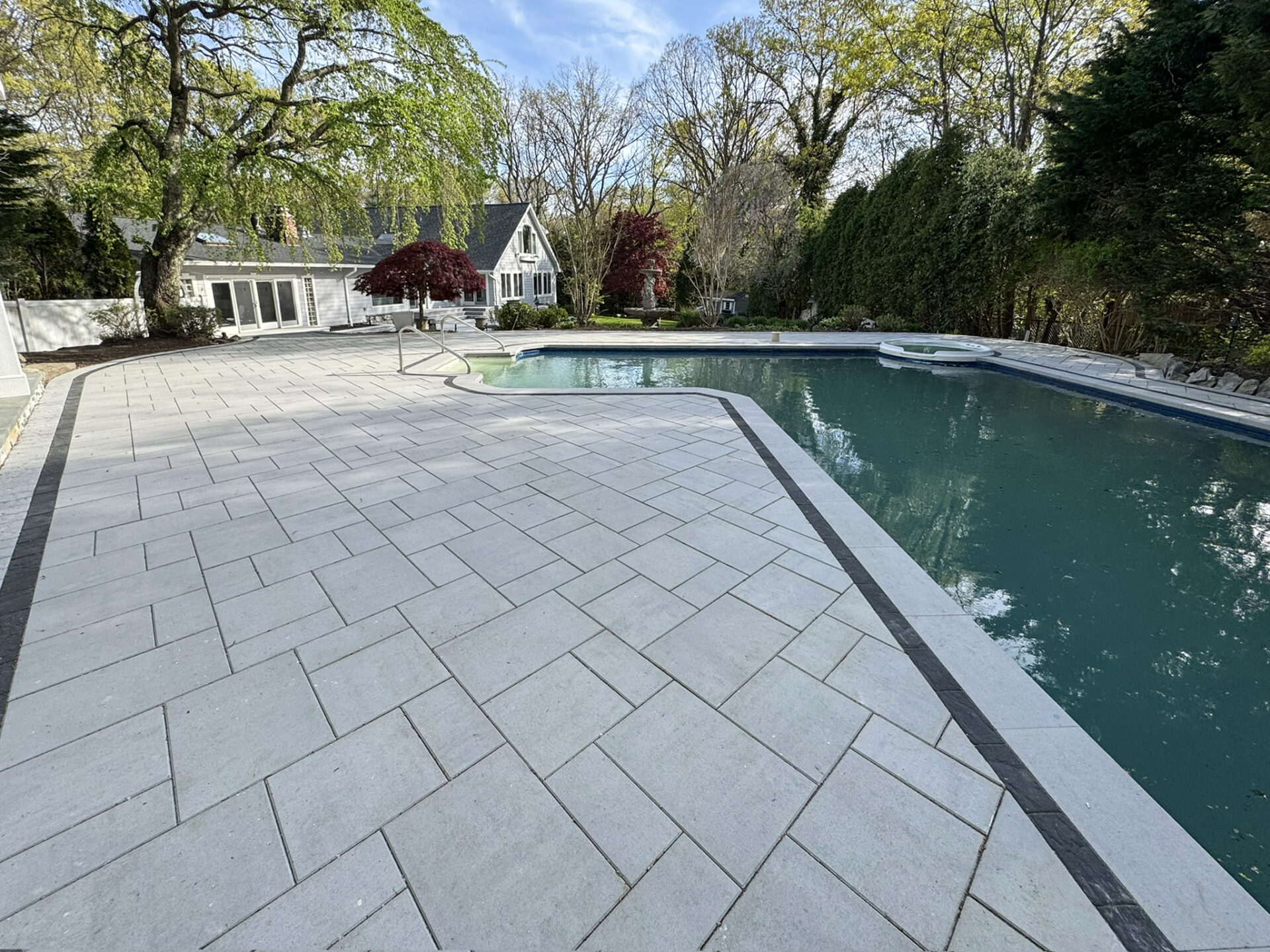 Large backyard with stone patio and swimming pool, surrounded by trees and shrubs, with a white house and clear sky in the background.