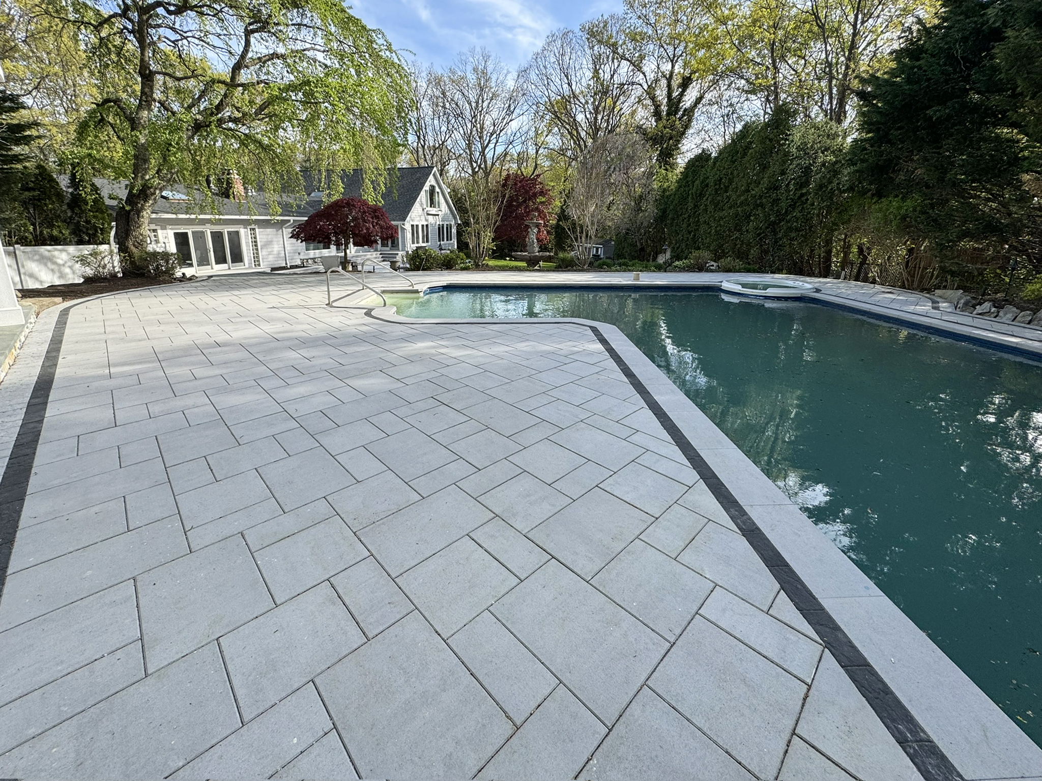 Large backyard with stone patio and swimming pool, surrounded by trees and shrubs, with a white house and clear sky in the background.