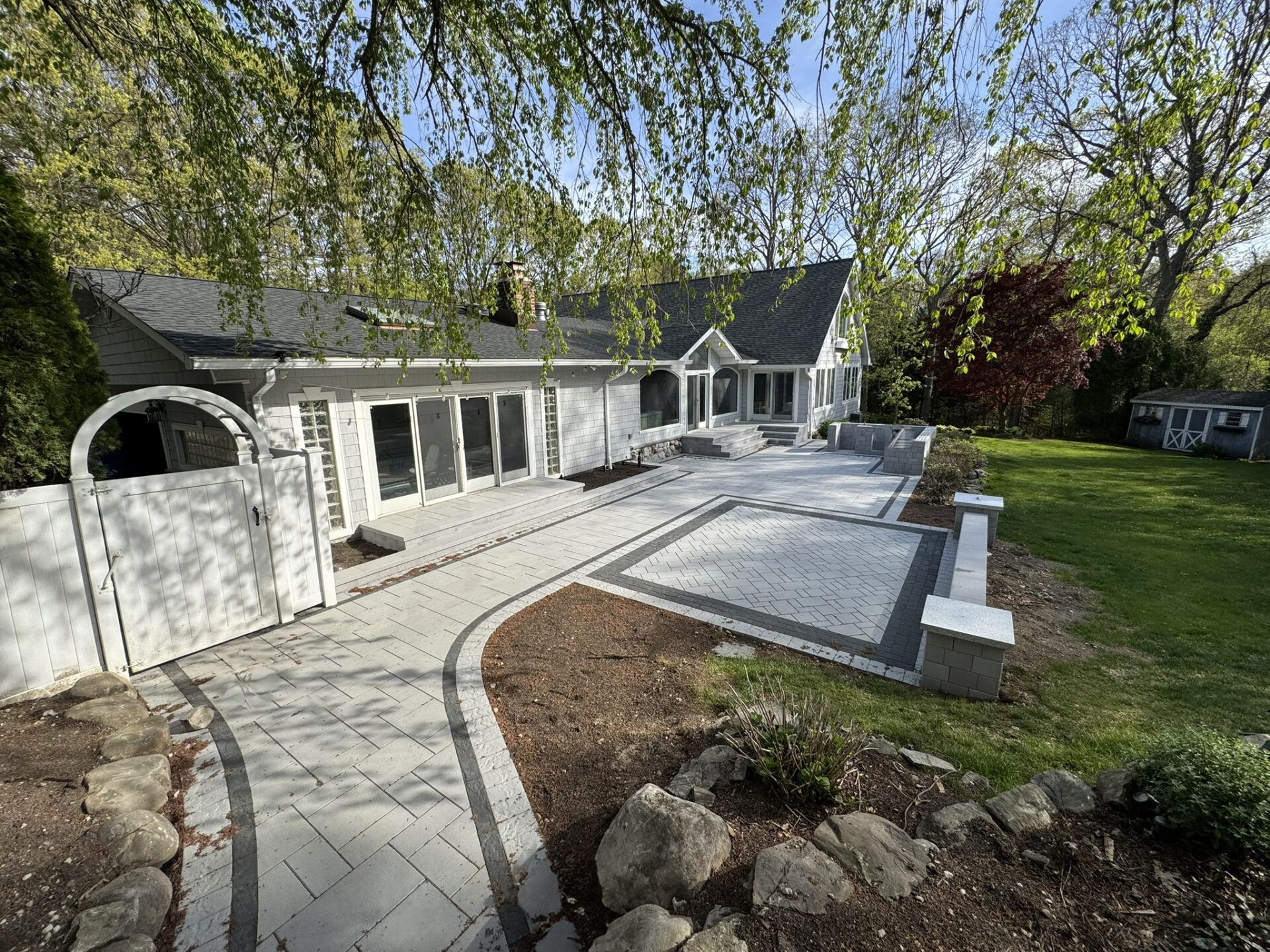 A white house with large windows overlooks a spacious stone patio with a walkway, surrounded by greenery and trees in a backyard.