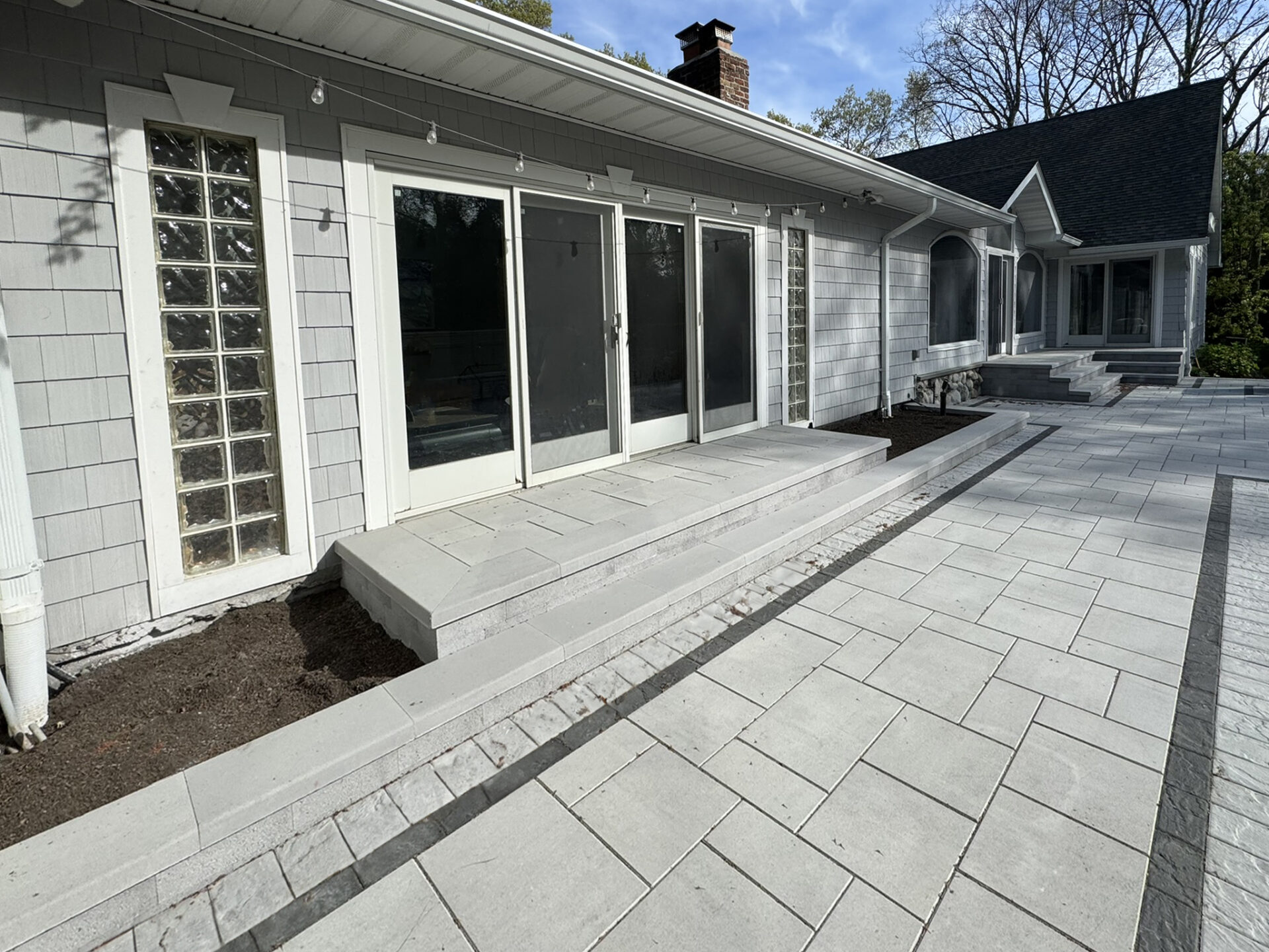 A modern patio with light gray stone pavers, wide steps, sliding glass doors, and a landscaped area along the house’s exterior.