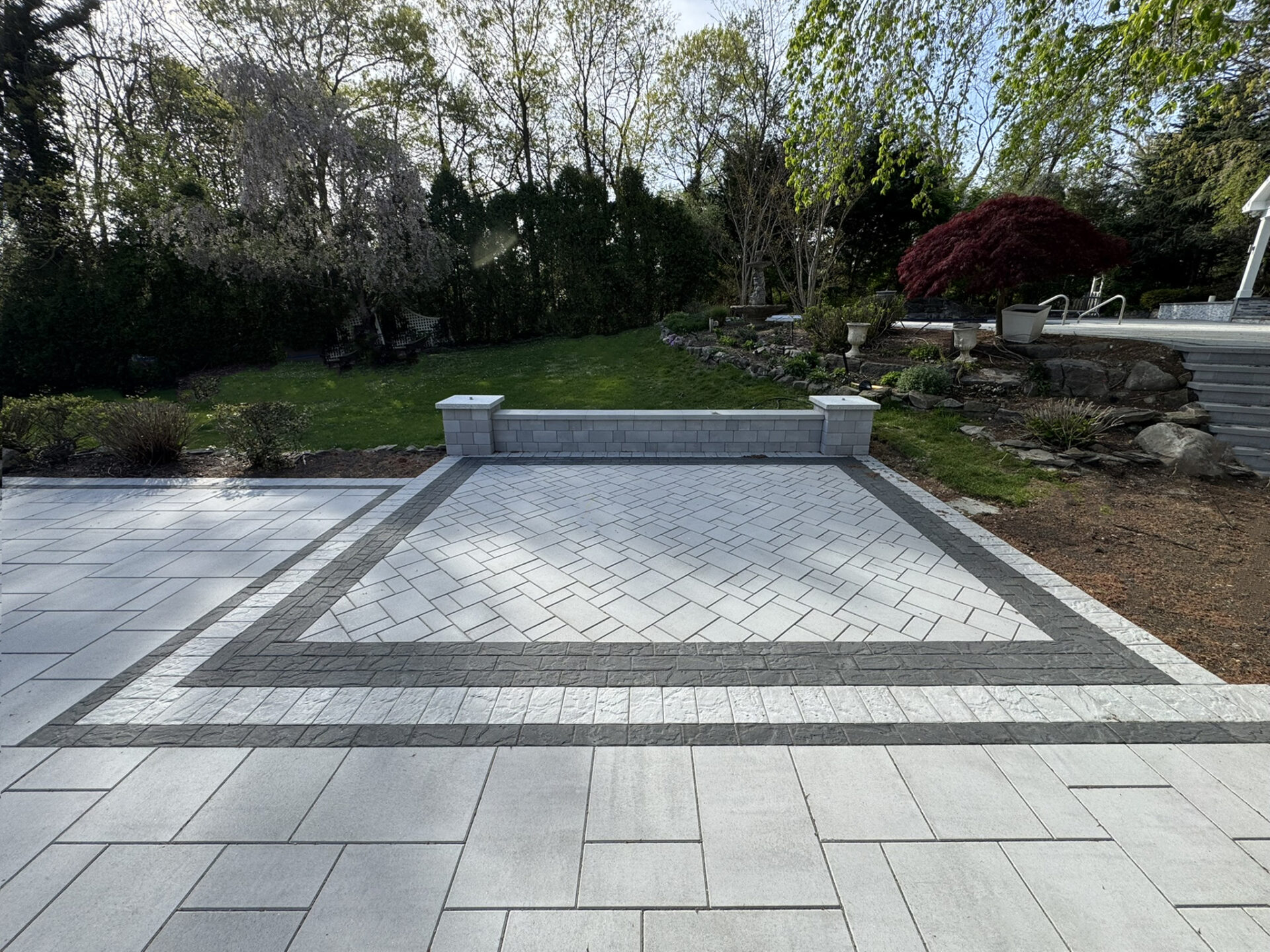 Rectangular paved patio area with a decorative stone border, low stone wall at the back, and landscaped yard with trees and shrubs in the background.