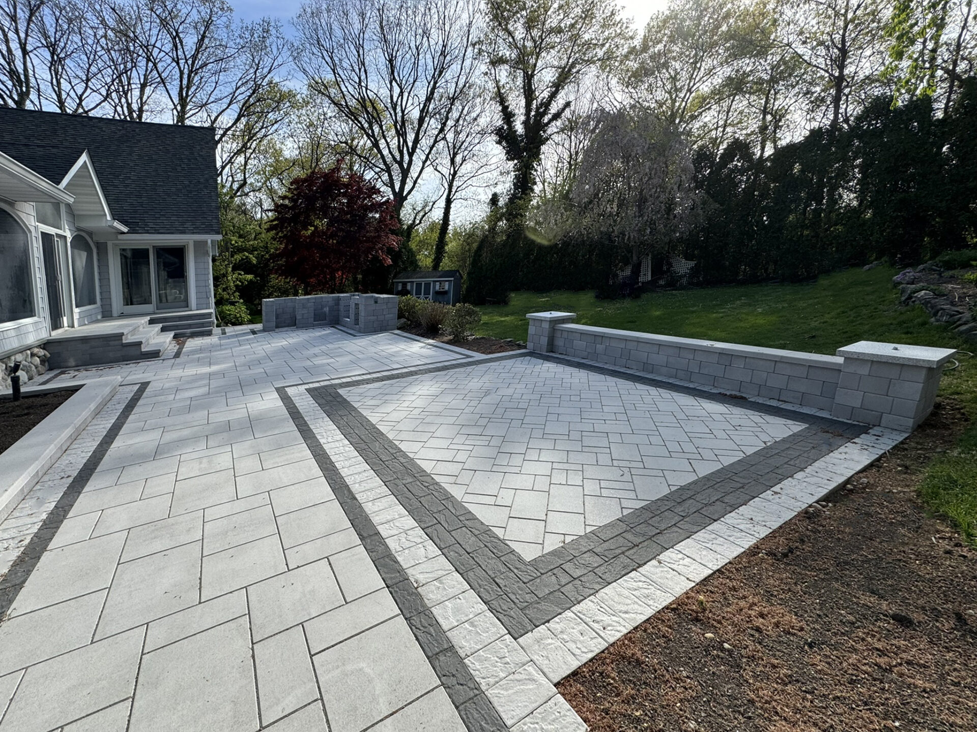 Large stone patio with geometric gray pavers, bordered by low stone walls, next to a house and lawn with trees in the background.