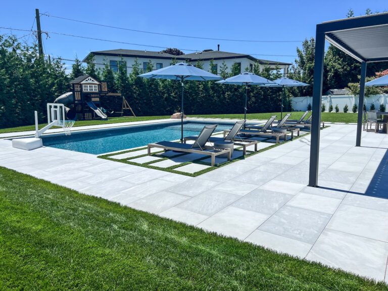A modern backyard features a rectangular pool, lounge chairs with umbrellas, a playset in the corner, and stone tile patio surrounded by grass.