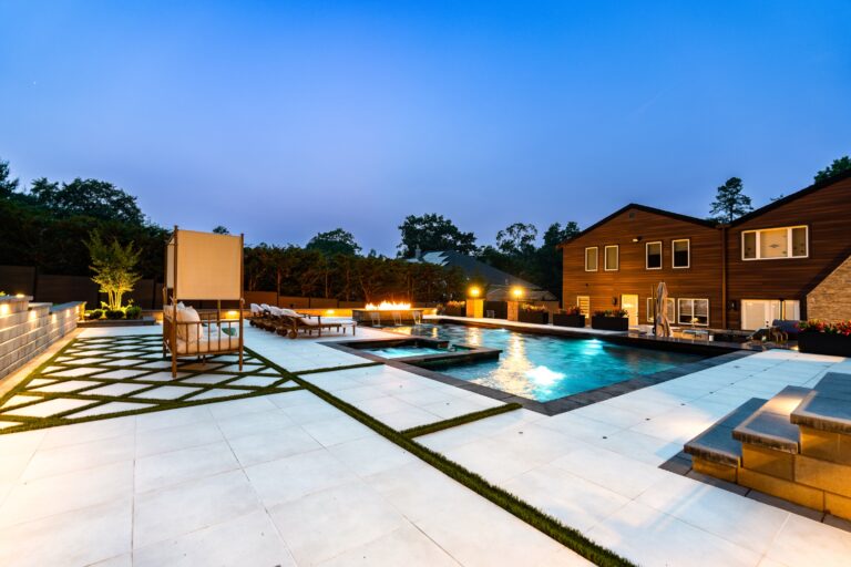 Modern backyard with a large swimming pool, fire pit, lounge chairs, tile flooring, and a contemporary house in the background at dusk.