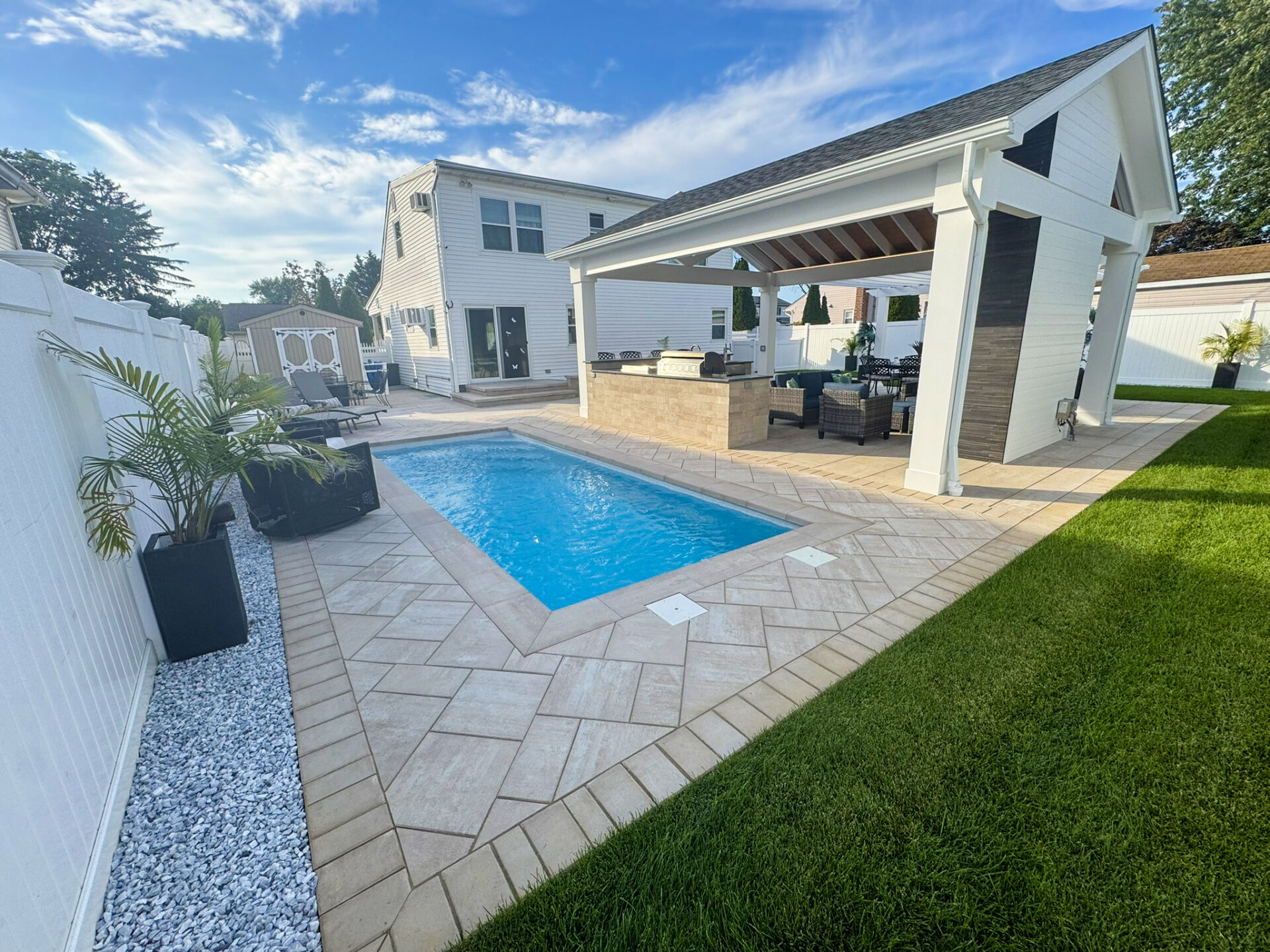 Rectangular backyard pool with lounge chairs, a covered patio area with seating, white fencing, and green lawn under a partly cloudy sky.