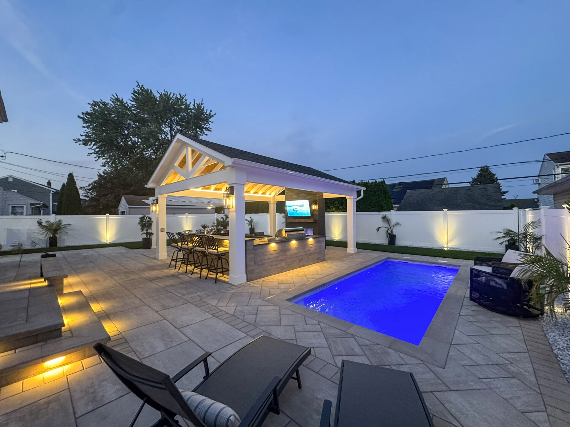 A modern backyard with a small illuminated pool, a covered outdoor bar and TV area, lounge chairs, and surrounding patio at dusk.