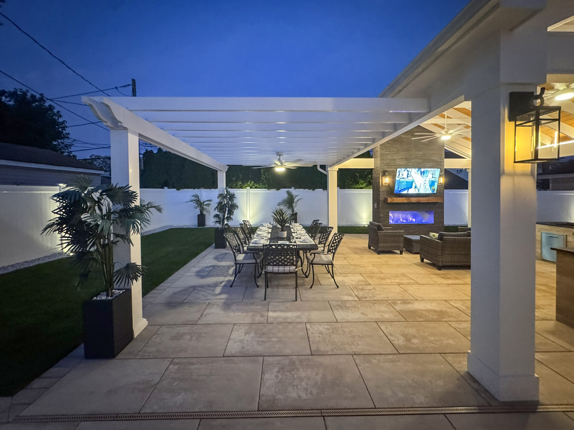 Covered outdoor patio at dusk with a dining table, chairs, potted plants, lounge area, TV, fireplace, and outdoor kitchen beside a lawn and white fence.