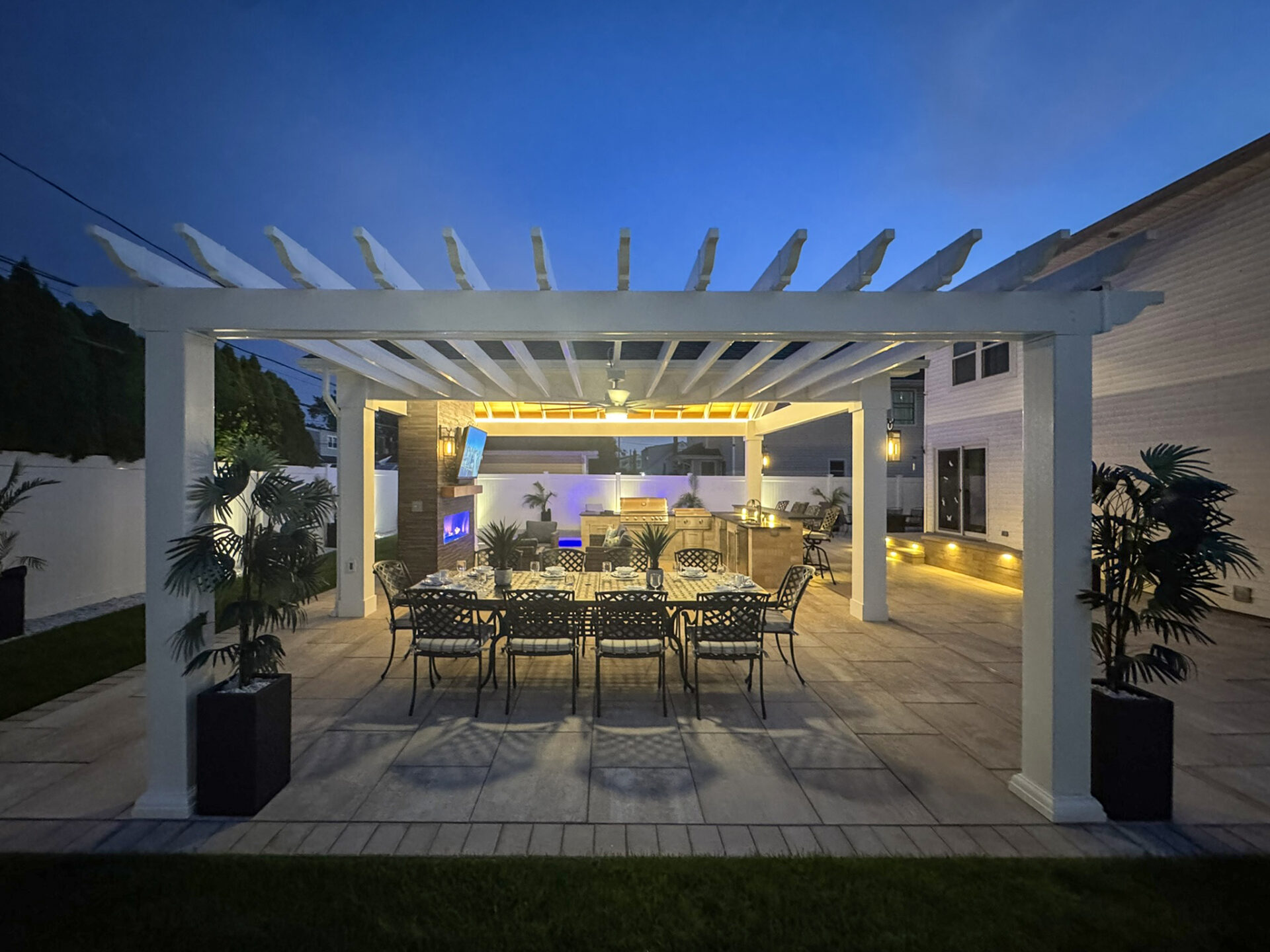 Outdoor patio with a white pergola, dining table and chairs, outdoor kitchen, potted plants, and ambient lighting at dusk.