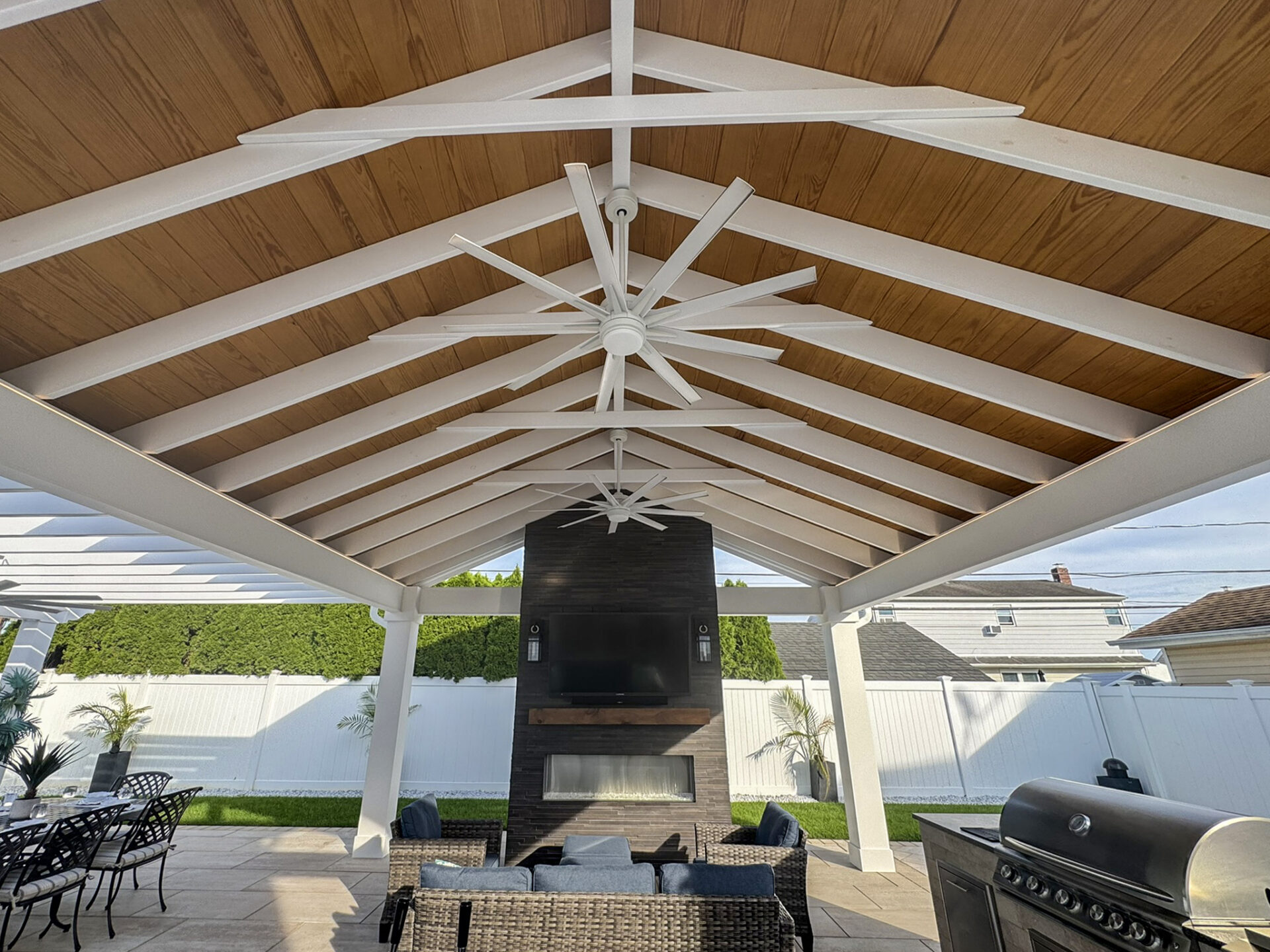 Covered patio with a wooden ceiling, white beams, two large ceiling fans, outdoor fireplace with a TV, wicker seating, and a grill area.