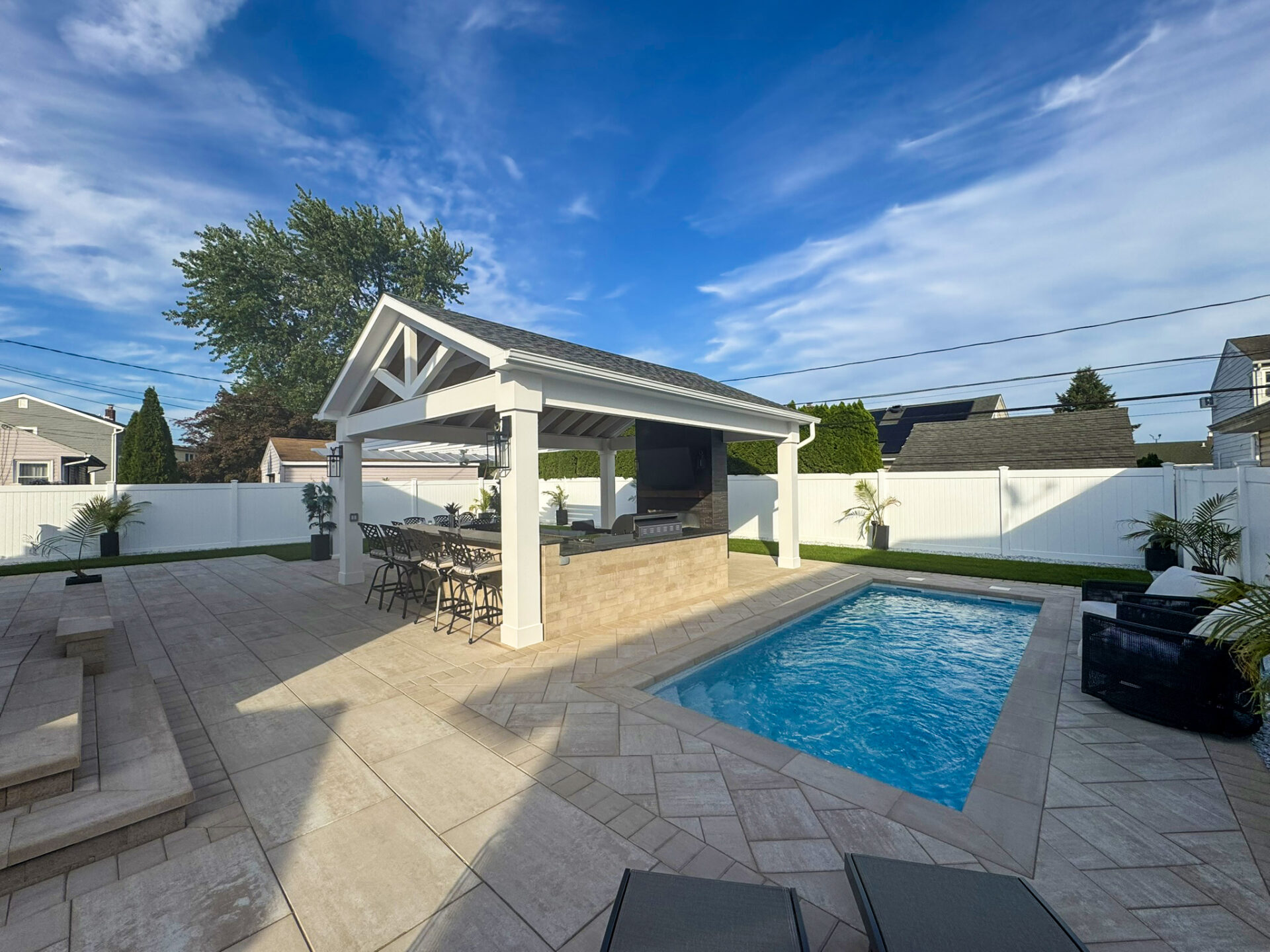 A backyard with a small rectangular pool, a covered outdoor bar area, lounge chairs, and a white fence under a blue sky.