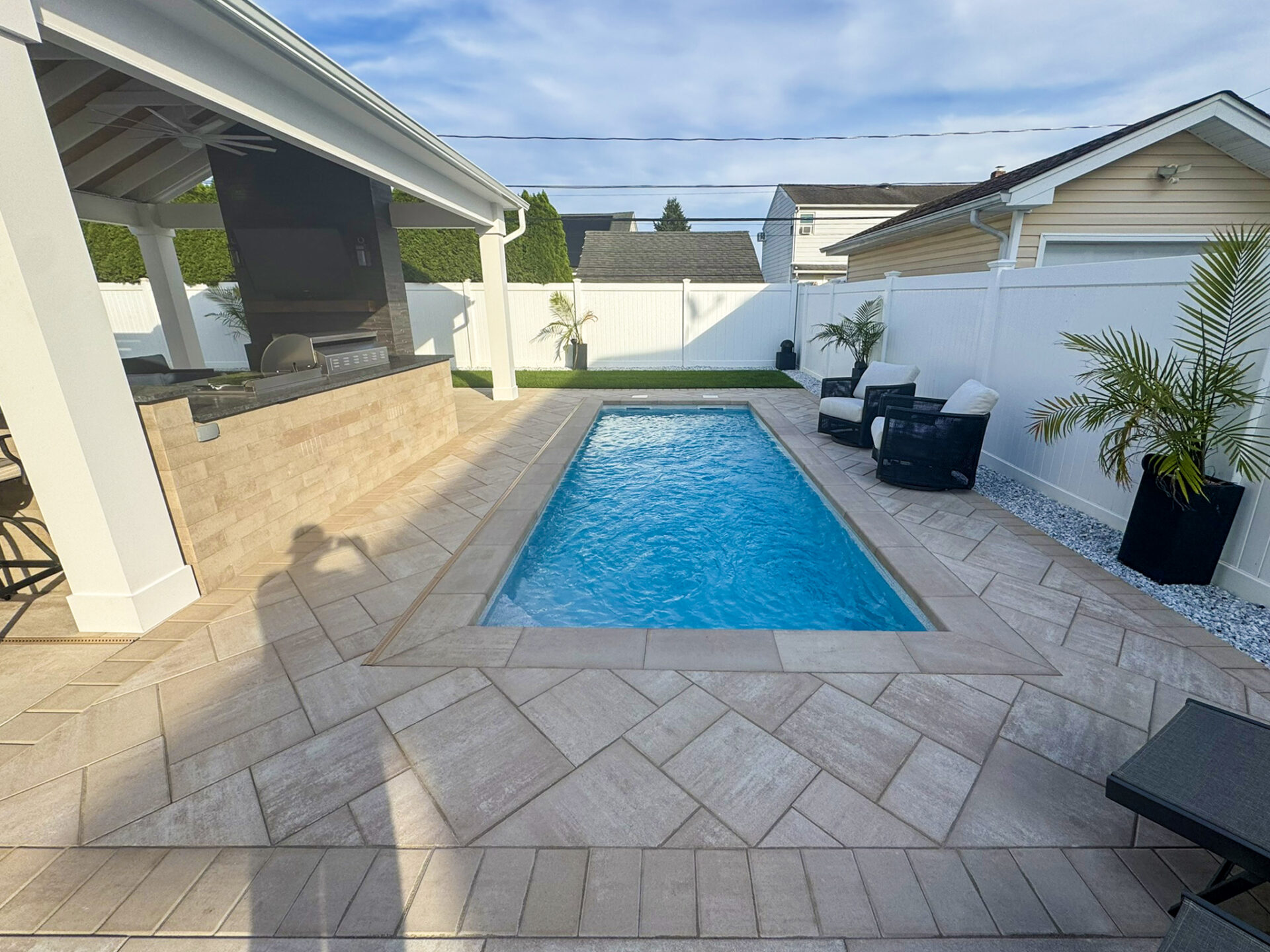 Rectangular backyard pool surrounded by tan pavers, patio seating, palm plants, and a covered outdoor kitchen area beside a white privacy fence.