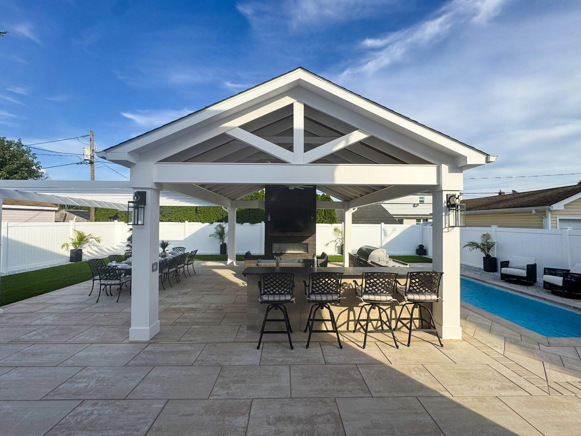 Covered outdoor patio with bar seating, dining tables, grill, and a pool, surrounded by white fencing and potted plants under a partly cloudy sky.
