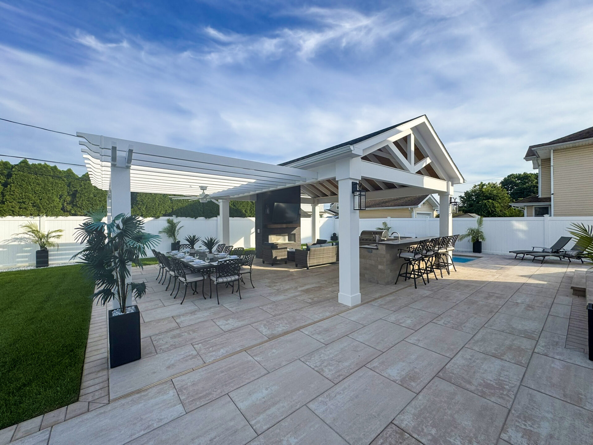 Outdoor patio with a covered dining area, bar, TV, lounge chairs, and potted plants on tiled flooring under a partly cloudy sky.
