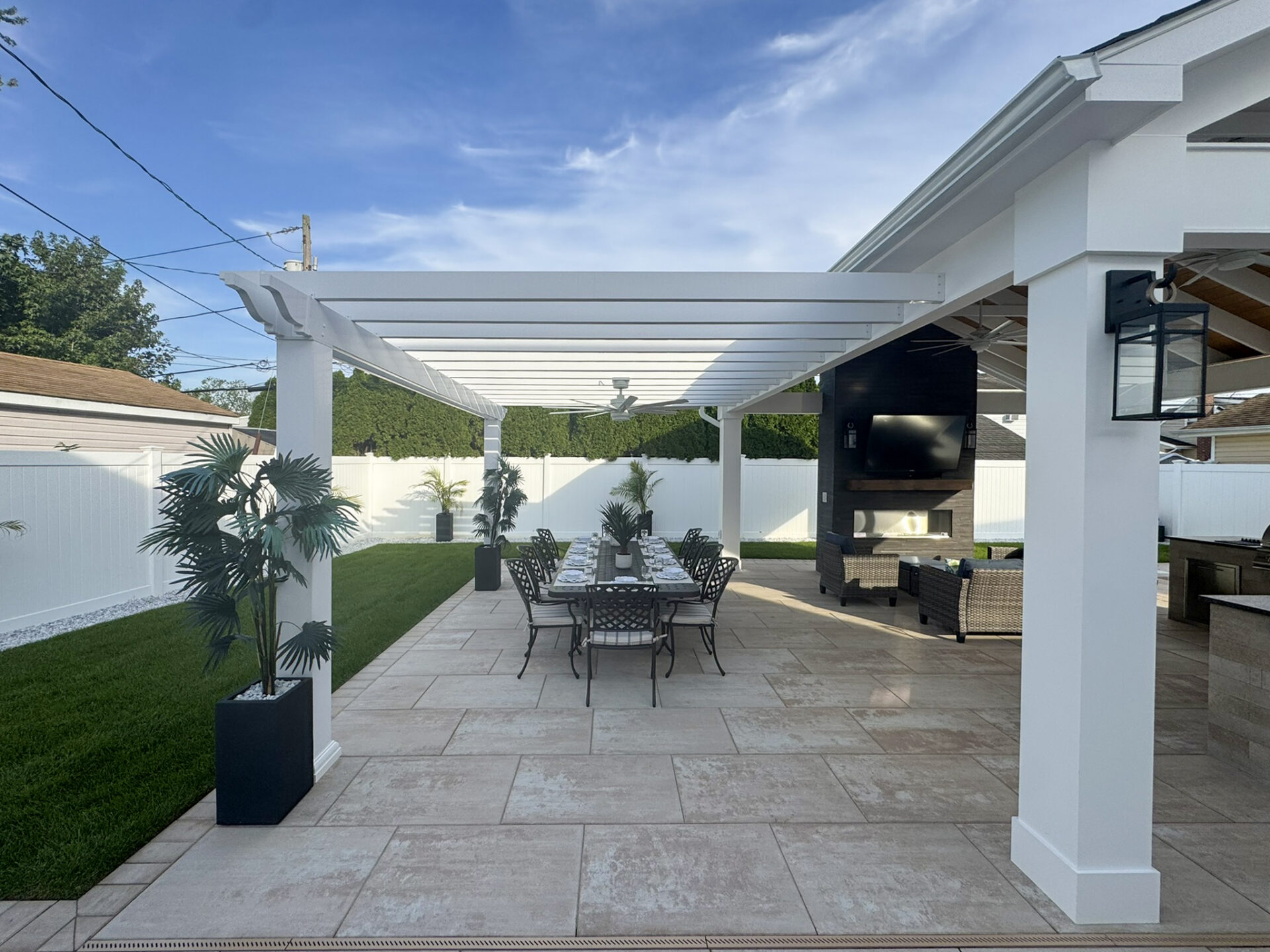 Outdoor patio with pergola, dining table and chairs, potted plants, tiled floor, and a seating area with a TV under a covered section, surrounded by a fenced yard.