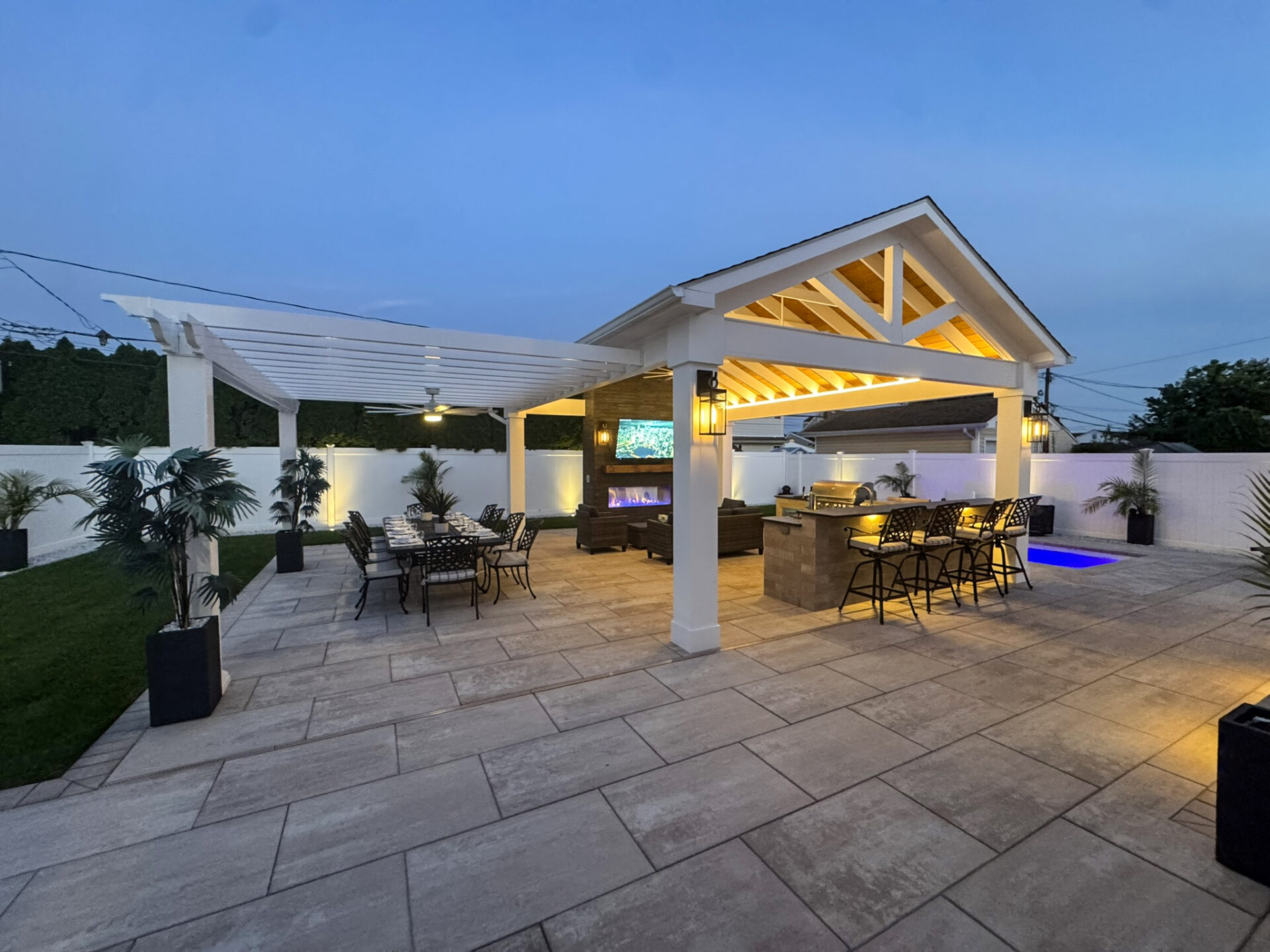 Outdoor patio with a covered seating area, dining table, bar, TV, and small pool, surrounded by potted plants and a white privacy fence at dusk.
