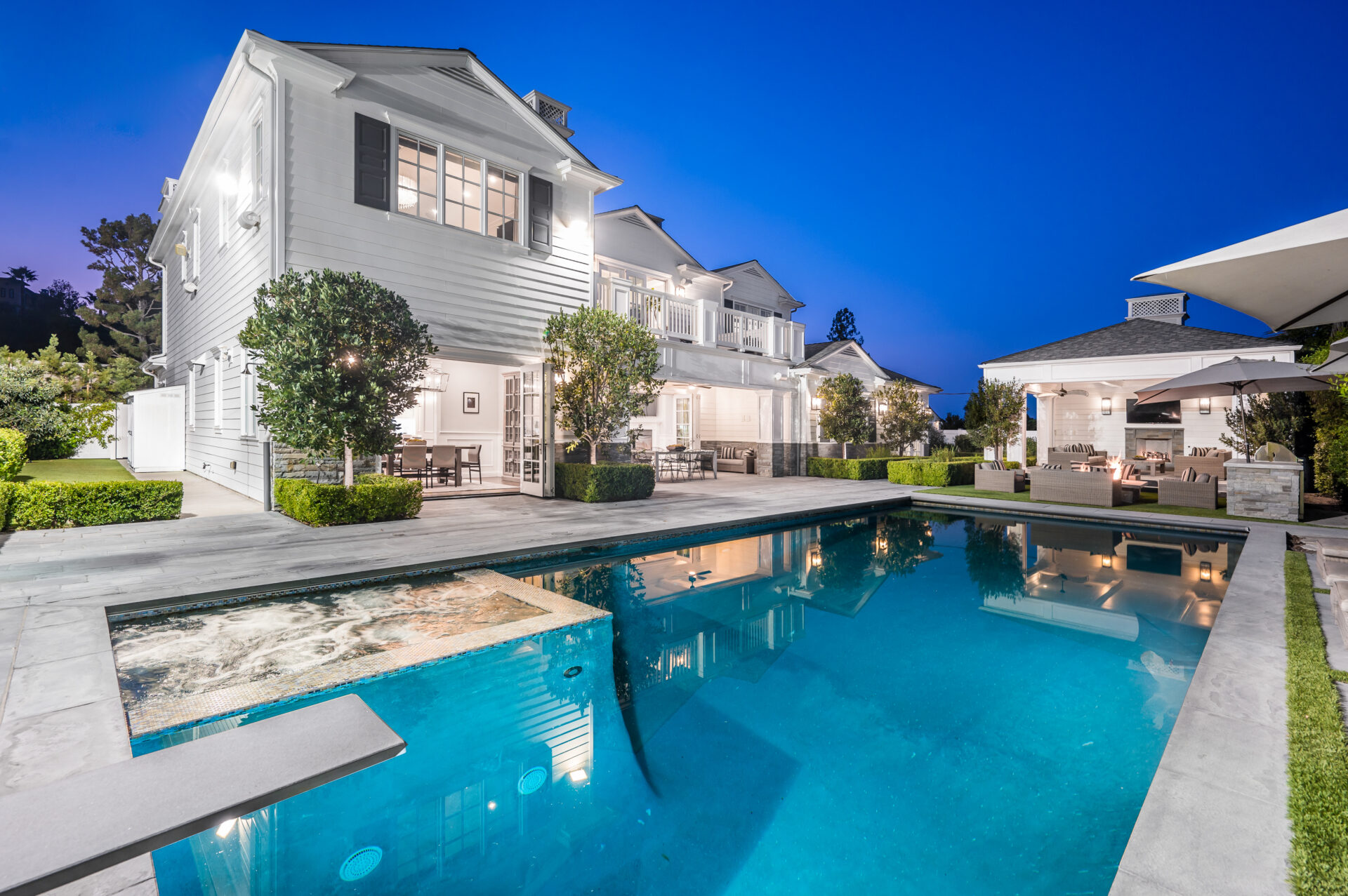 Modern two-story white house with large windows, pool, hot tub, poolside patio, and outdoor seating area, photographed at dusk with lights on.
