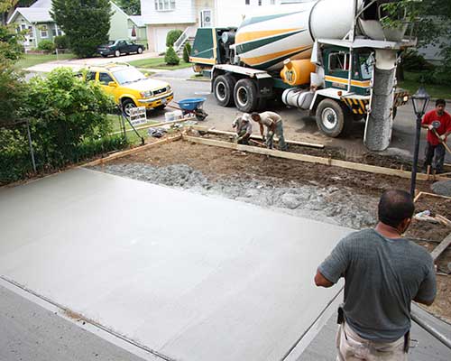 Workers pouring and smoothing cement for a residential driveway with a concrete mixer truck and other tools in the background.