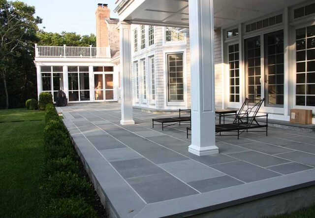 Elegant home exterior with a spacious patio, white pillars, and large windows surrounded by lush greenery.