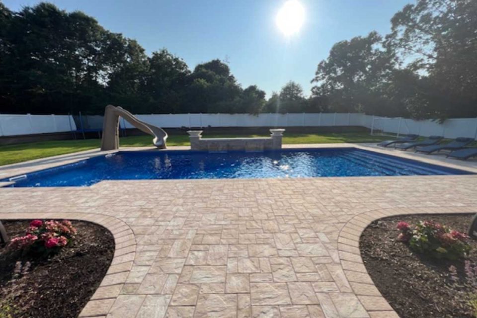 A sunlit backyard featuring a clear blue swimming pool with a slide and a stone patio surrounded by a white fence.