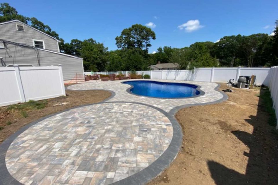 A residential backyard featuring a blue kidney-shaped pool, surrounded by a newly paved patio with a winding pathway, bordered by a white fence under a clear sky.