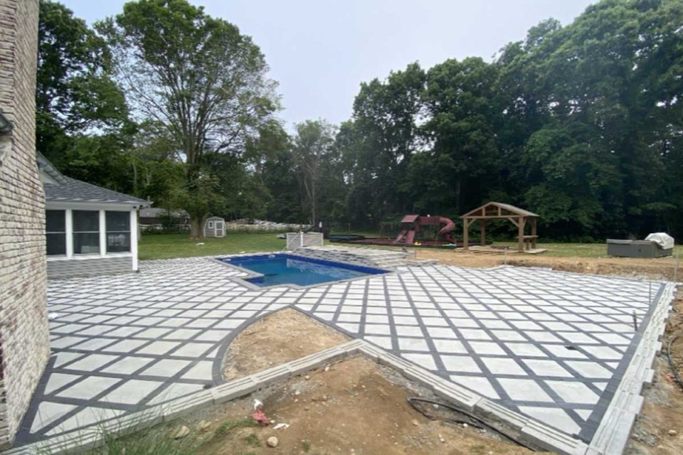 A residential backyard under construction with a patterned patio surrounding an in-ground pool, a playground in the distance, and surrounded by trees.