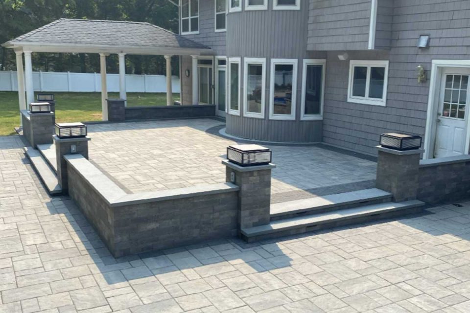 Backyard of a house with a large stone patio, a few steps, and built-in lighting on stone posts, featuring a covered gazebo in the background.