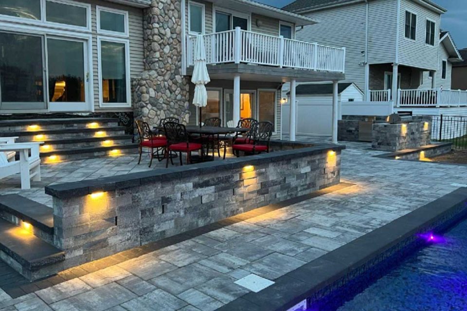 A well-lit backyard at dusk with stone patio steps, a pool, and elegant seating areas next to a large house.