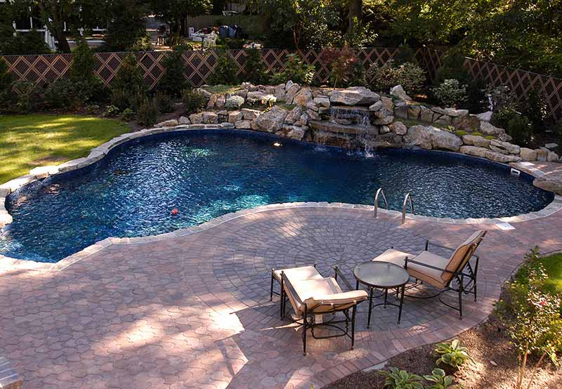 A kidney-shaped swimming pool with a small waterfall, surrounded by a paved patio with chairs and tables, enclosed by a fenced garden.