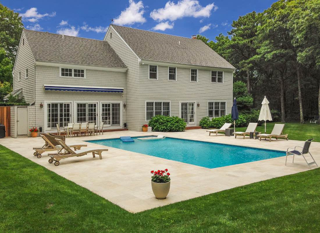 A large two-story gray house with a blue-striped awning, surrounded by green trees, featuring a backyard with a pool, sun loungers, and a patio area.