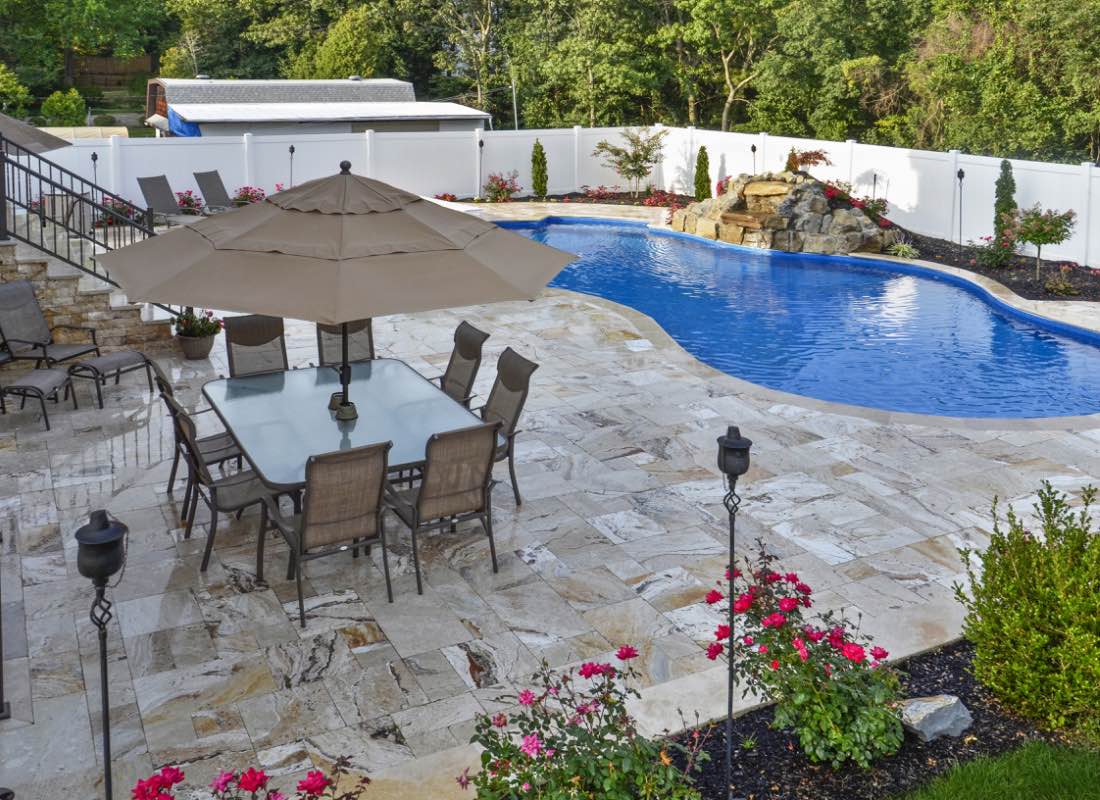 A backyard featuring a blue swimming pool with a stone deck, patio dining set under an umbrella, surrounding greenery, and a white fence.