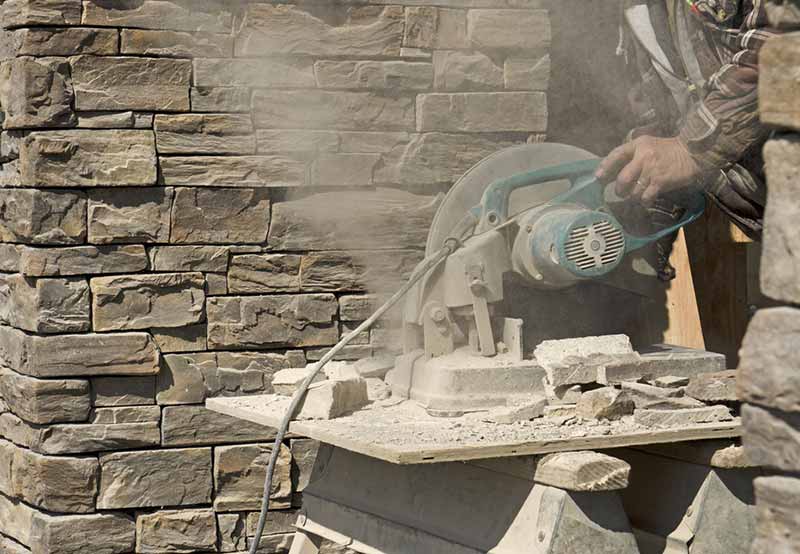 A person using a circular saw to cut stone, generating a cloud of dust, with a stone wall in the background.