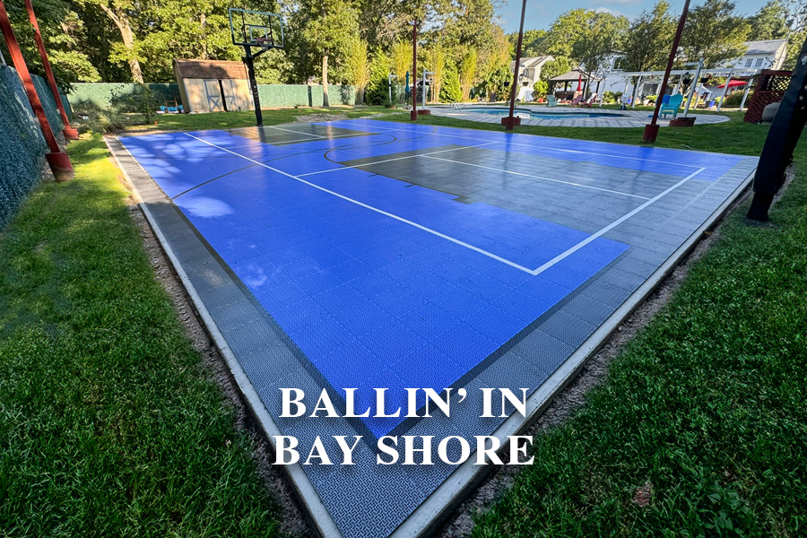 Outdoor basketball court with blue and gray surface, surrounded by grass, trees, and a playground area in the background. Text reads "BALLIN’ IN BAY SHORE.