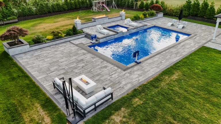 A rectangular backyard pool with two fire features, nearby lounge chairs, a fire pit with seating, and a children's playset surrounded by greenery.