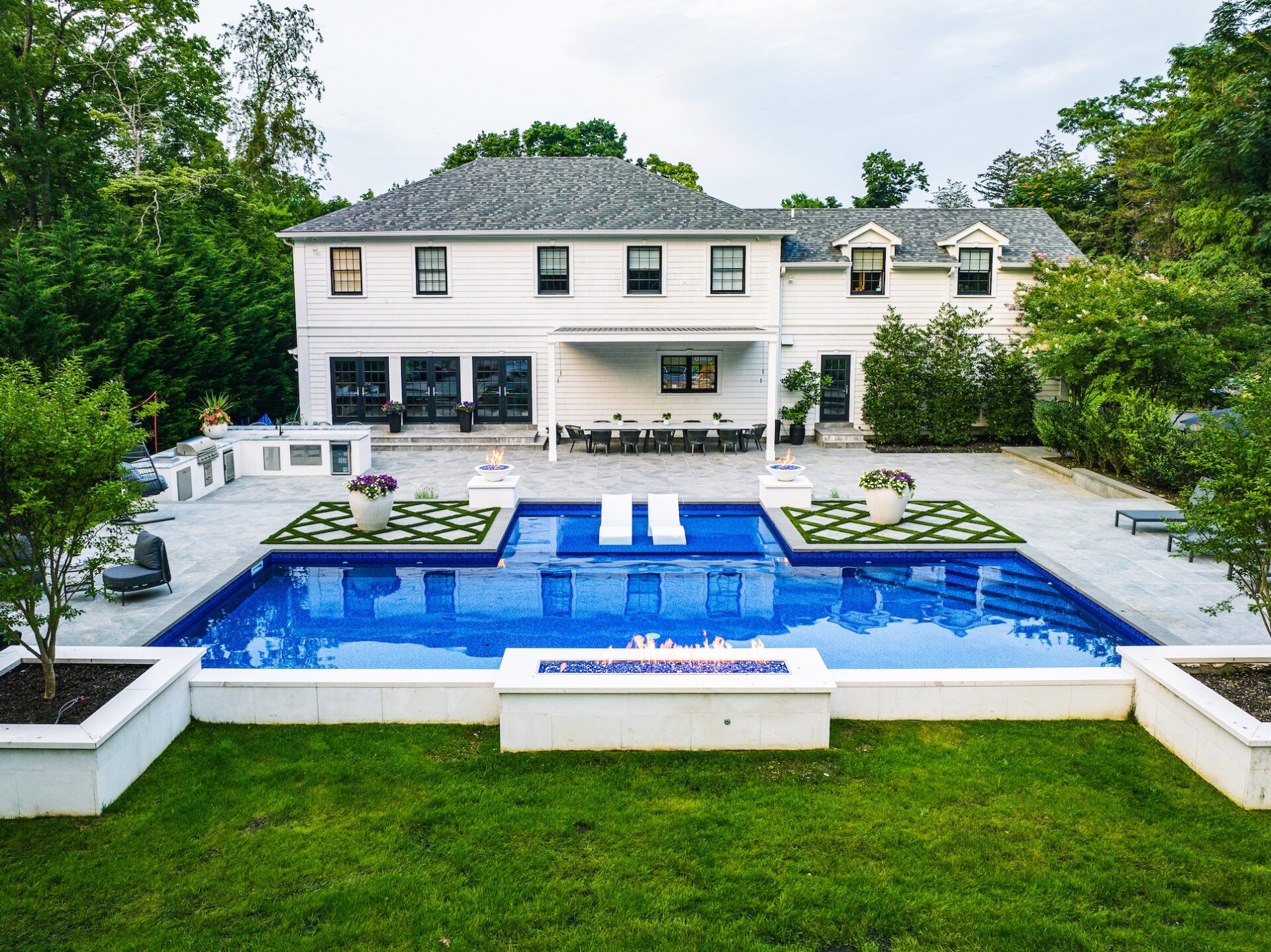 A modern backyard with a rectangular pool, sun loungers, manicured lawn, and a large white house in the background.