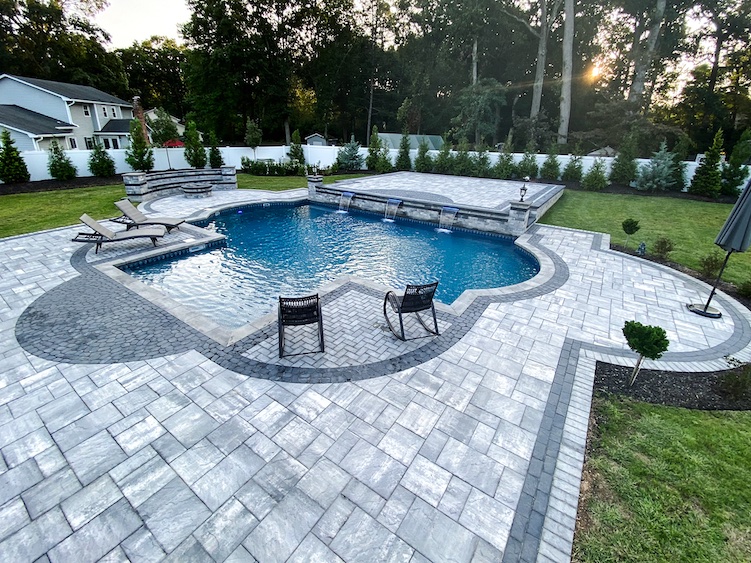 Rectangular outdoor swimming pool with attached spa, surrounded by gray stone patio, lounge chairs, wicker chairs, and landscaped yard with trees and a white fence in the background.