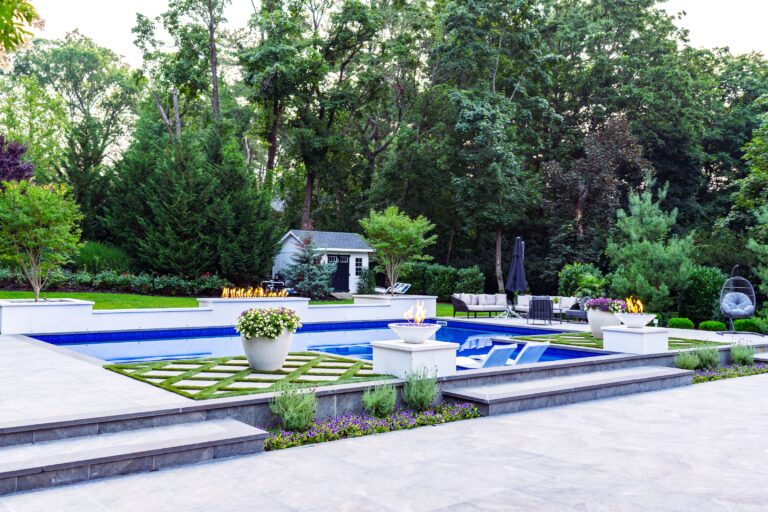 Modern backyard with a rectangular pool, in-pool lounge chairs, fire features, large planters, manicured lawn, and a small shed surrounded by trees and greenery.